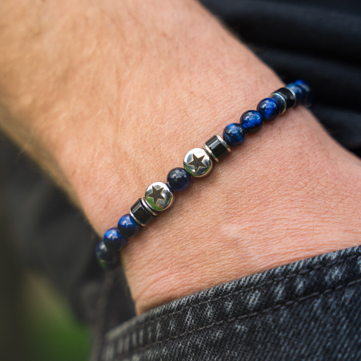Bracciale artigianale da uomo indossato. Realizzato con occhio di tigre blu, agata nera e acciaio sul quale sono incise due stelle. Lab93Studio
