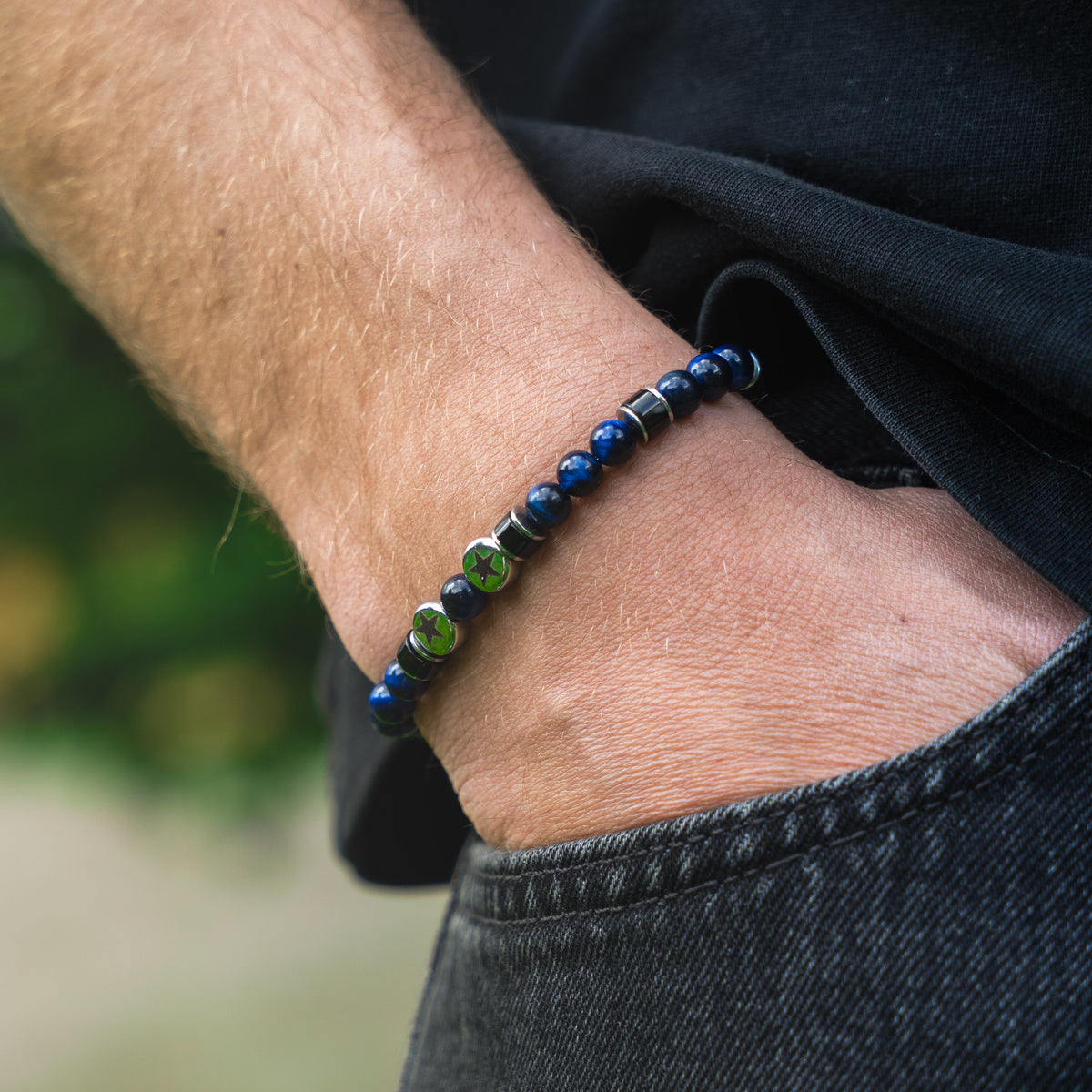 Bracciale artigianale da uomo indossato. Realizzato con occhio di tigre blu, agata nera e acciaio sul quale sono incise due stelle. Lab93Studio